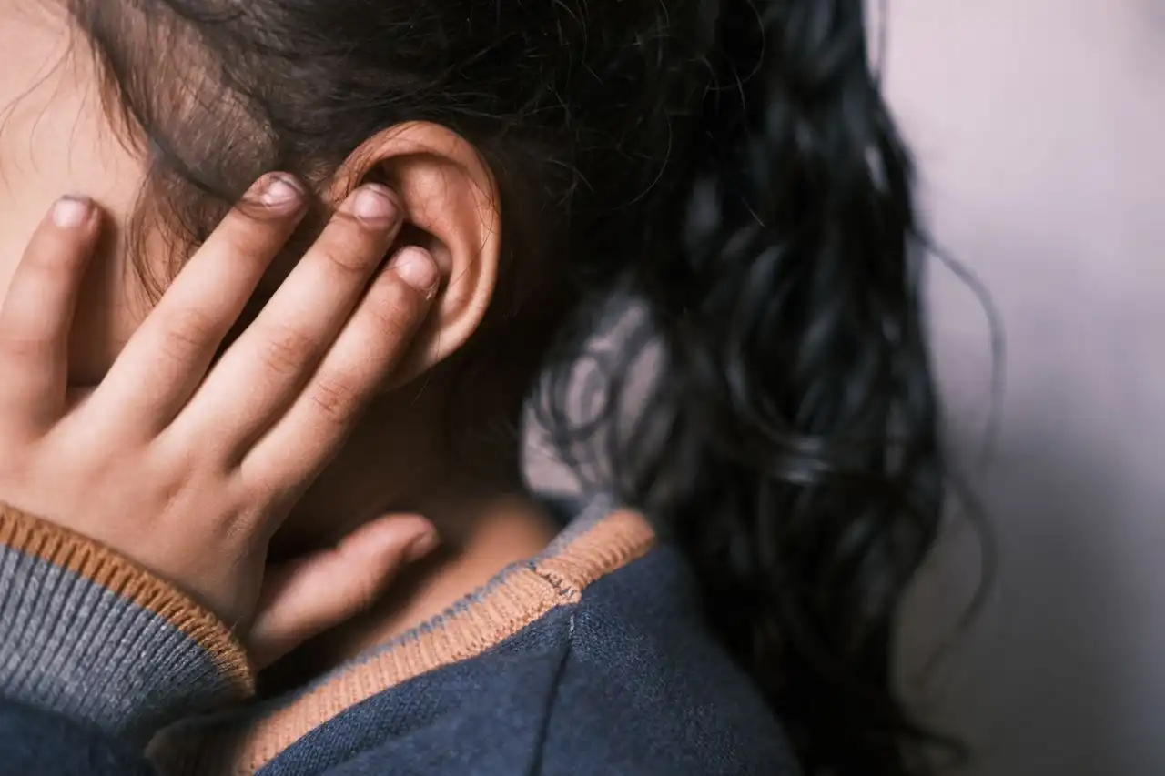 Close-up from behind of a child with long dark hair pressing their fingers to the side of the head next to one ear